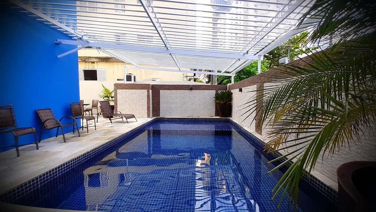 Piscina com azulejos azuis, sob uma cobertura branca. Cadeiras à esquerda; uma parede com padrão texturizado ao fundo.