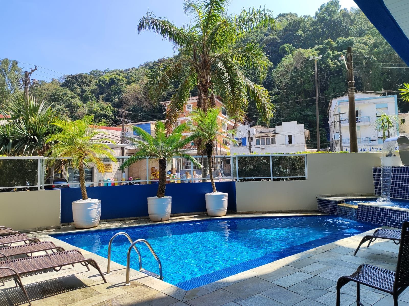 Piscina com palmeiras, água azul e prédios ao fundo. Dia ensolarado.