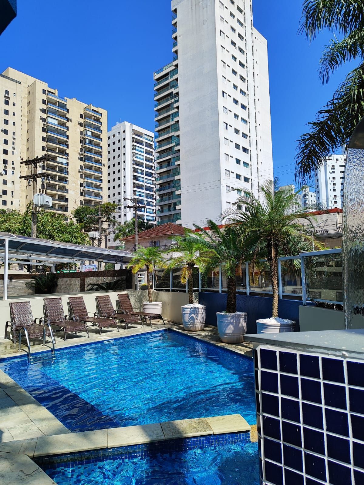 Uma piscina azul cercada por espreguiçadeiras e palmeiras, com altos prédios de apartamentos ao fundo.