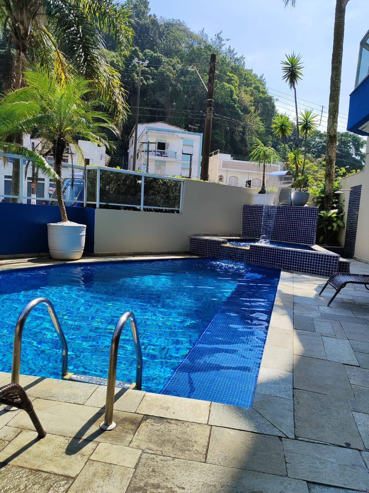 Piscina com água azul e degraus, cercada por um pátio claro e uma pequena cachoeira. Árvores verdes e prédios brancos ao fundo.