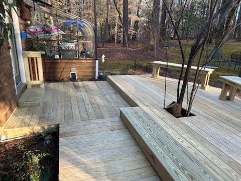 Wooden deck with built-in benches surrounding a tree, greenhouse in the background, surrounded by woods.
