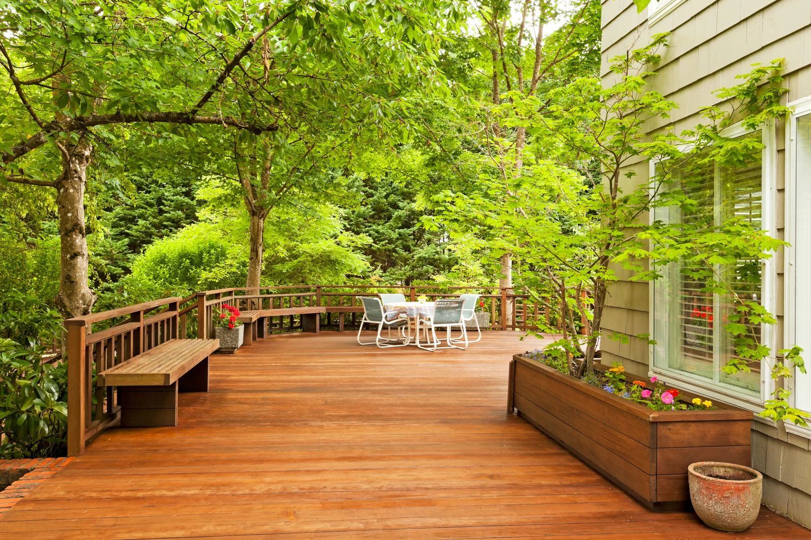 Wooden deck with benches and table set under trees, next to a house with a window.