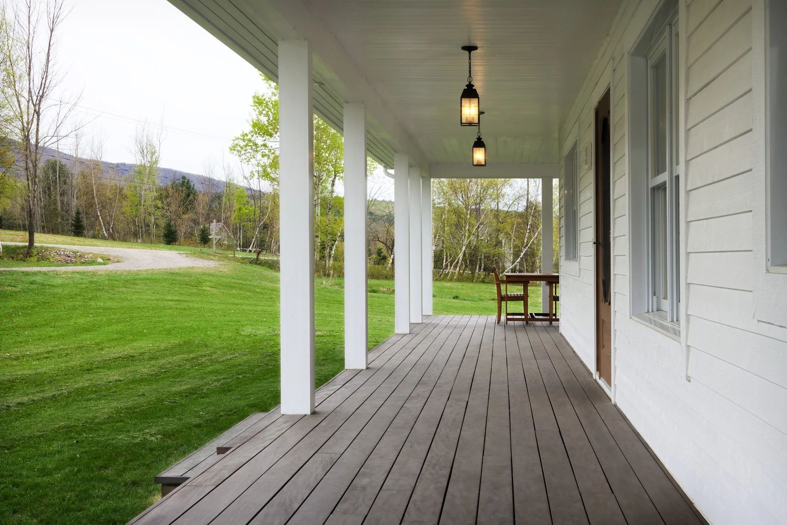 Covered farmhouse porch with white columns, wood deck, hanging lights, and grassy yard view