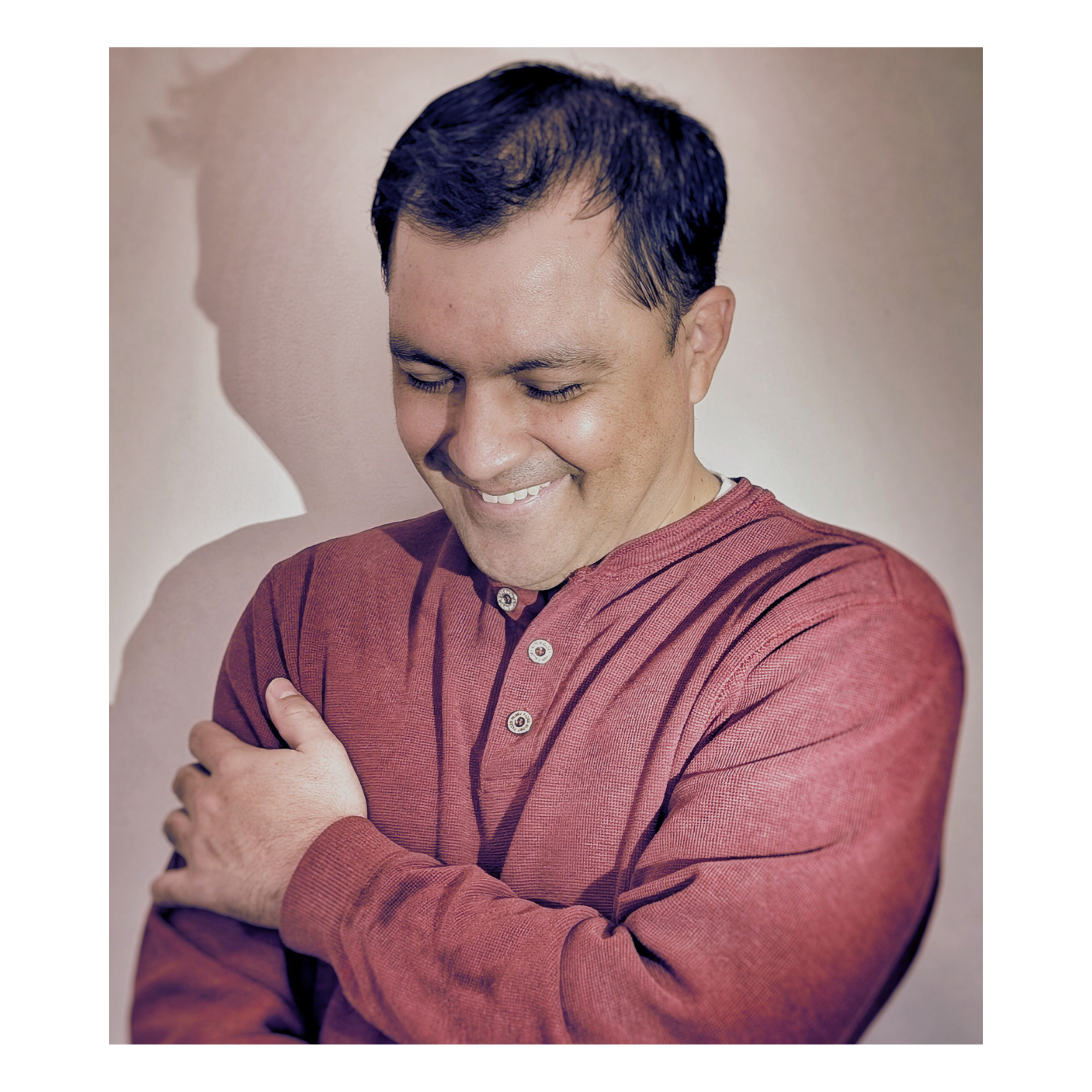 Smiling person in a red shirt looking down with arms crossed against a light background