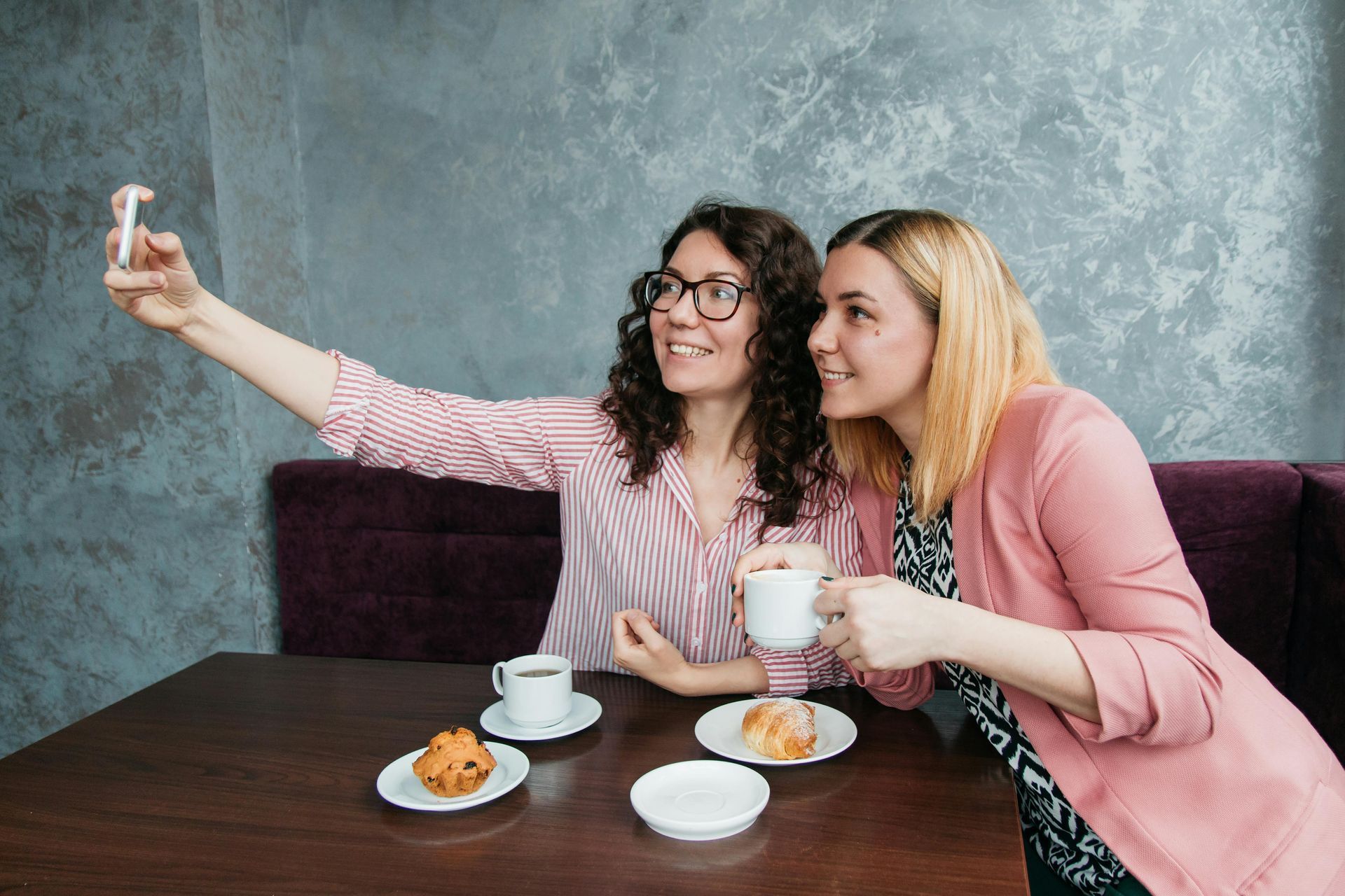 Two women taking a selfie in a cafe, smiling. One holds a phone, the other a mug. Baked goods on table.
