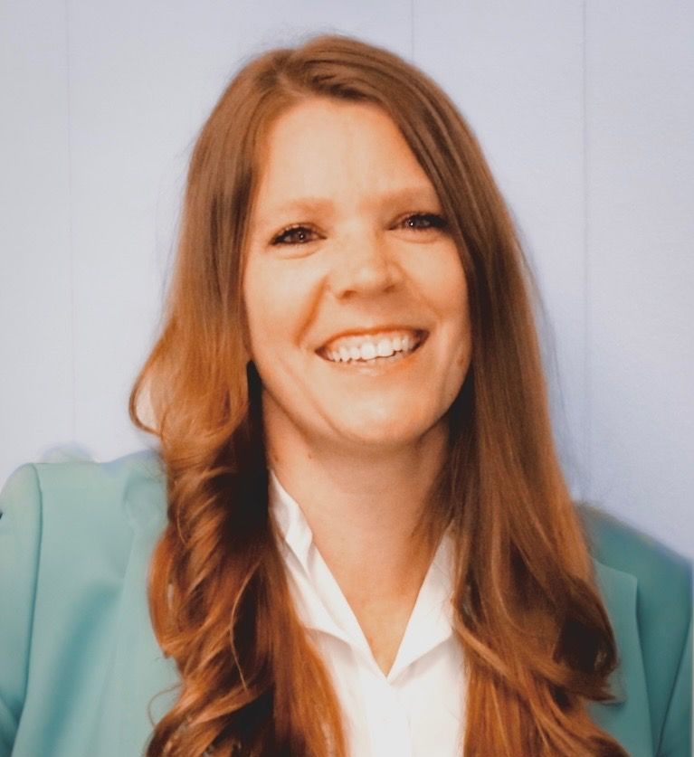 Smiling woman with long red hair in a teal blazer and white blouse against a light blue background