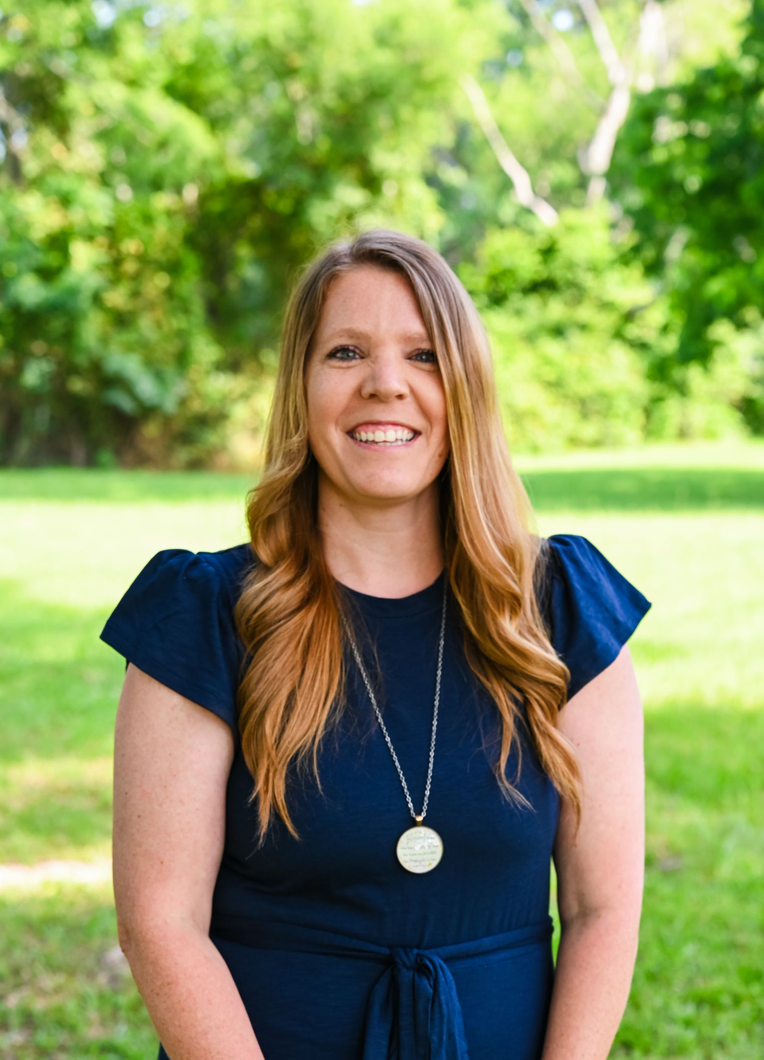 Smiling woman in a navy dress standing outdoors with green trees in the background