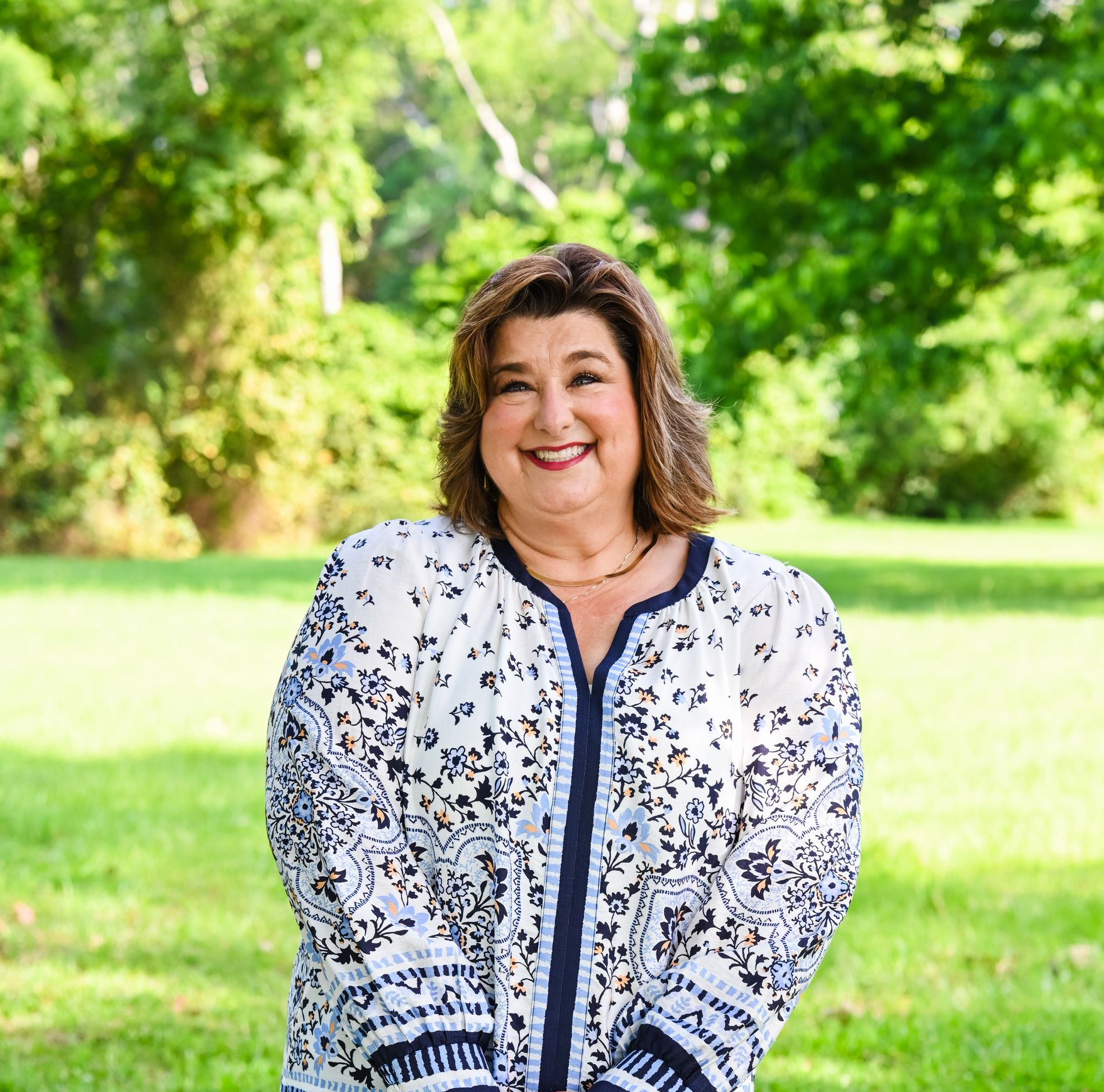 Smiling woman in a white patterned blouse standing outdoors in a green park