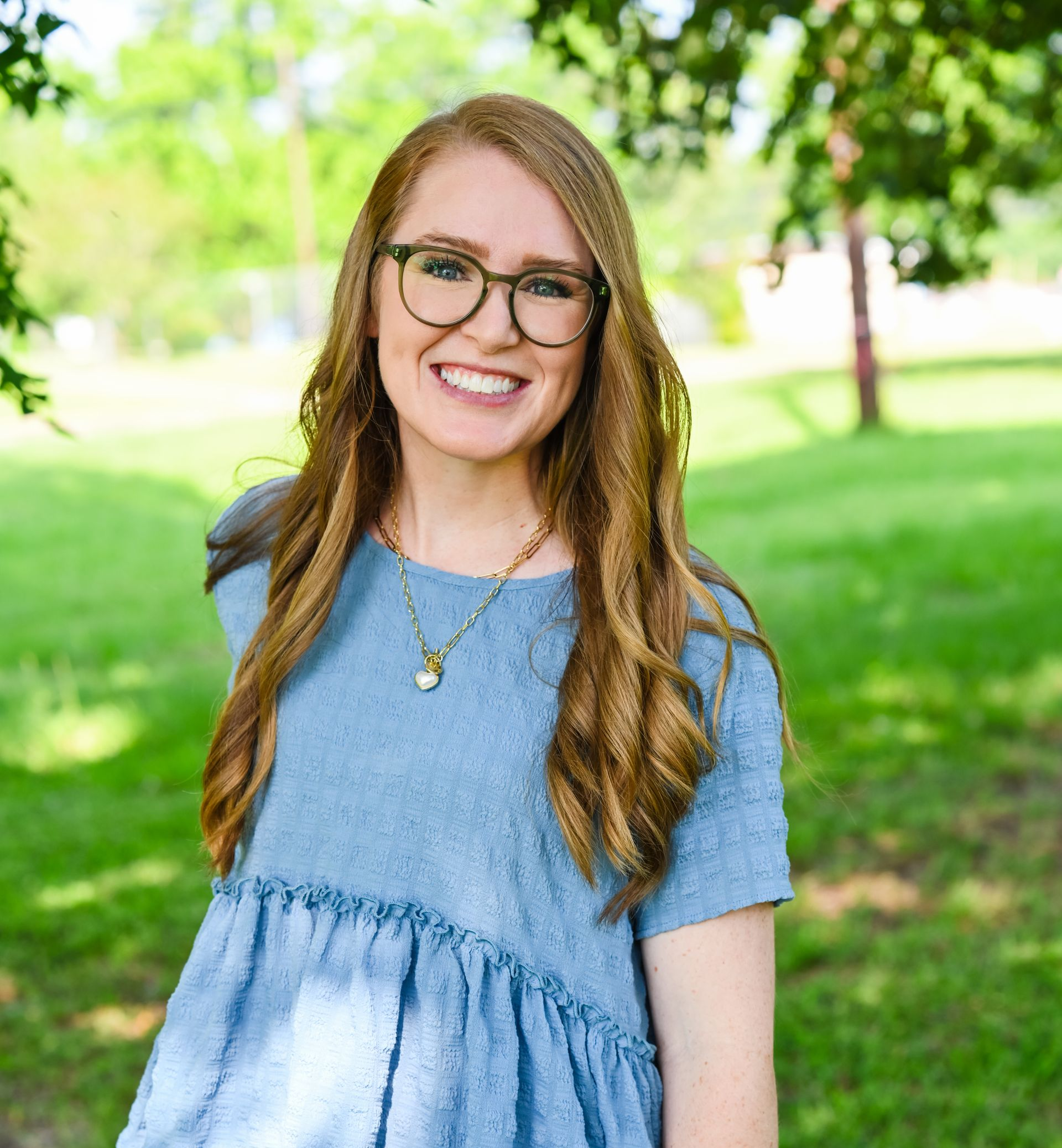Smiling person with glasses in a blue dress standing outdoors in a green park