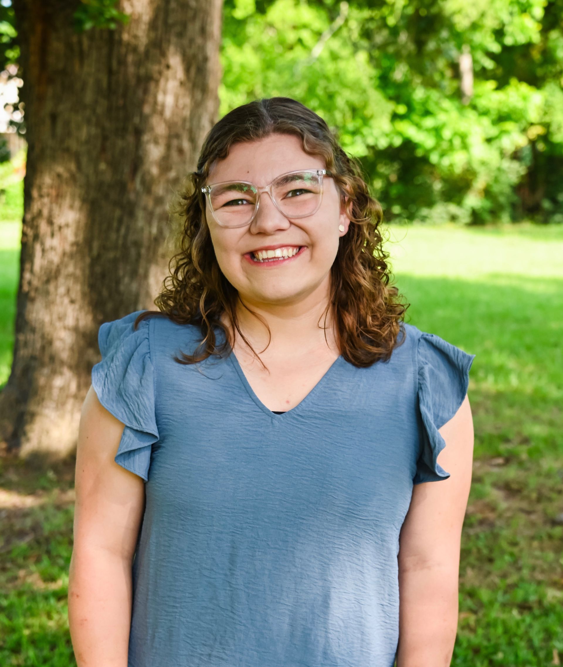 Smiling person in glasses and a blue top standing outdoors in a green park area