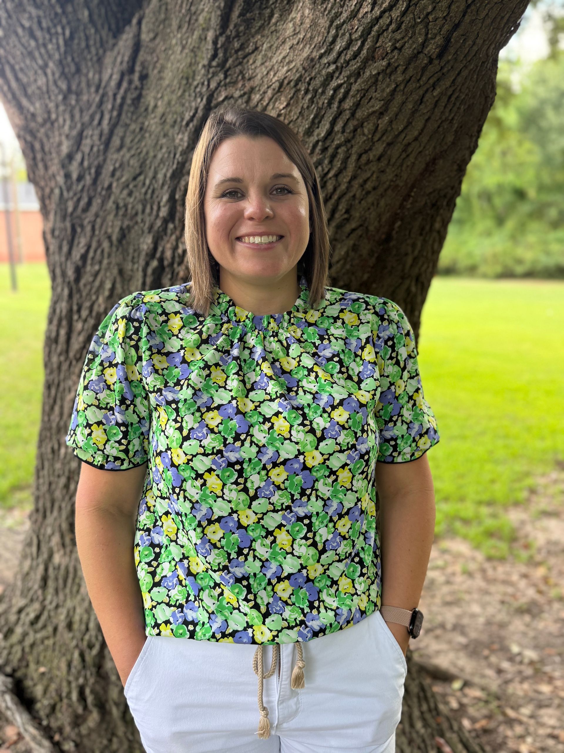 Smiling person in glasses and blue blouse standing outdoors near a tree and green lawn