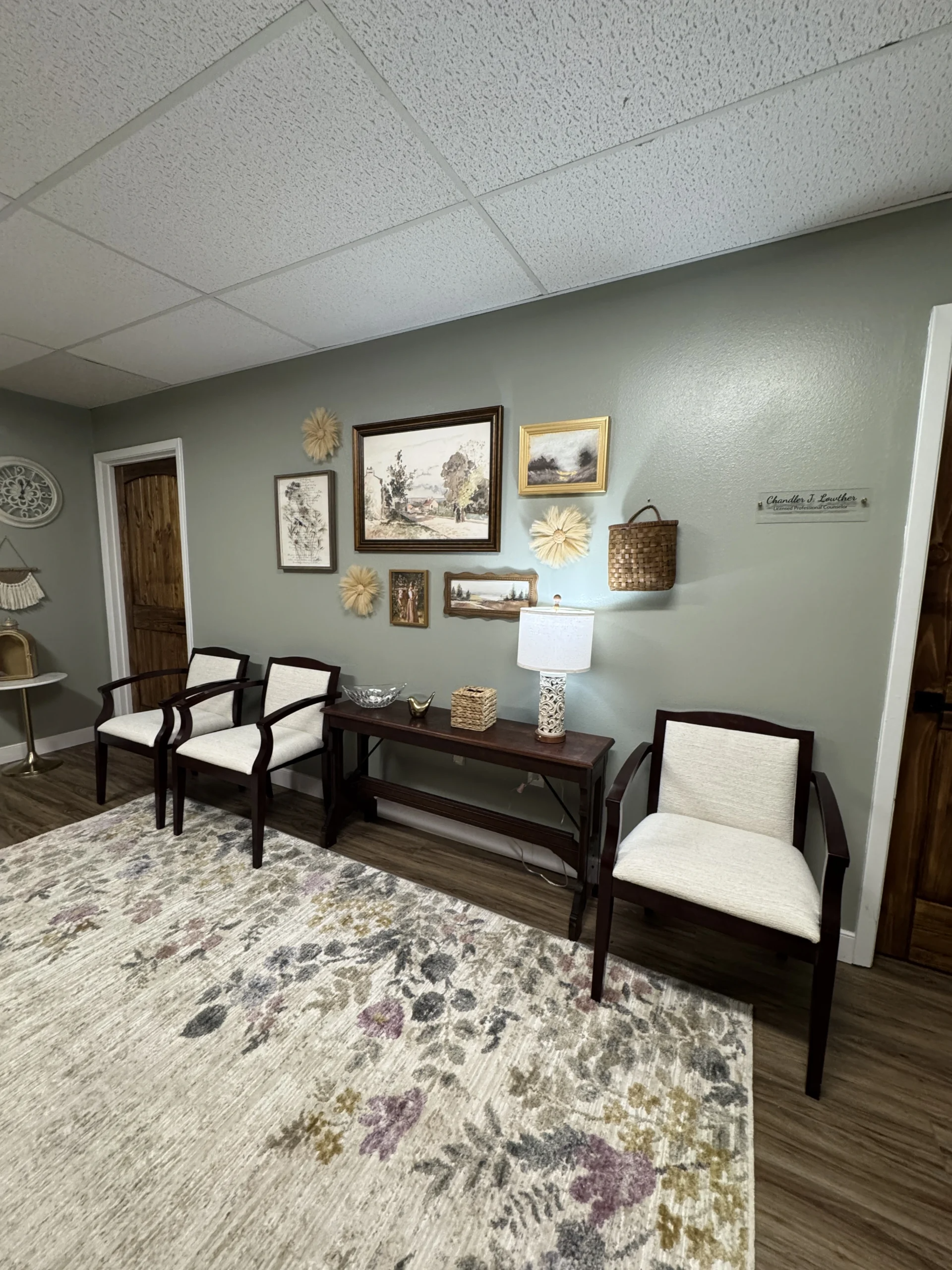 Waiting room with white chairs, dark bench, patterned rug, and framed wall art on gray walls.