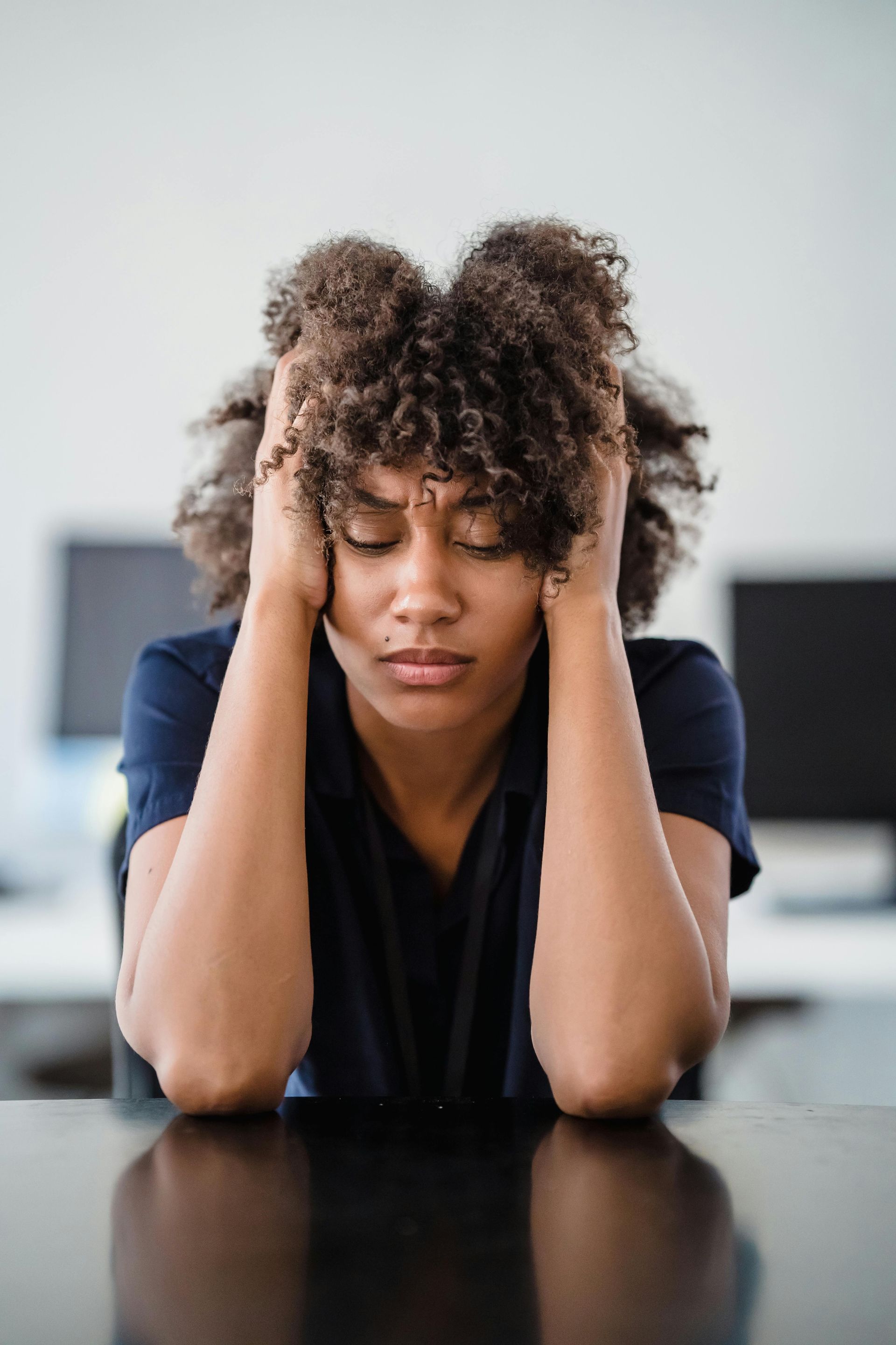 Person sitting at a table, resting their head in their hands with eyes closed, looking stressed