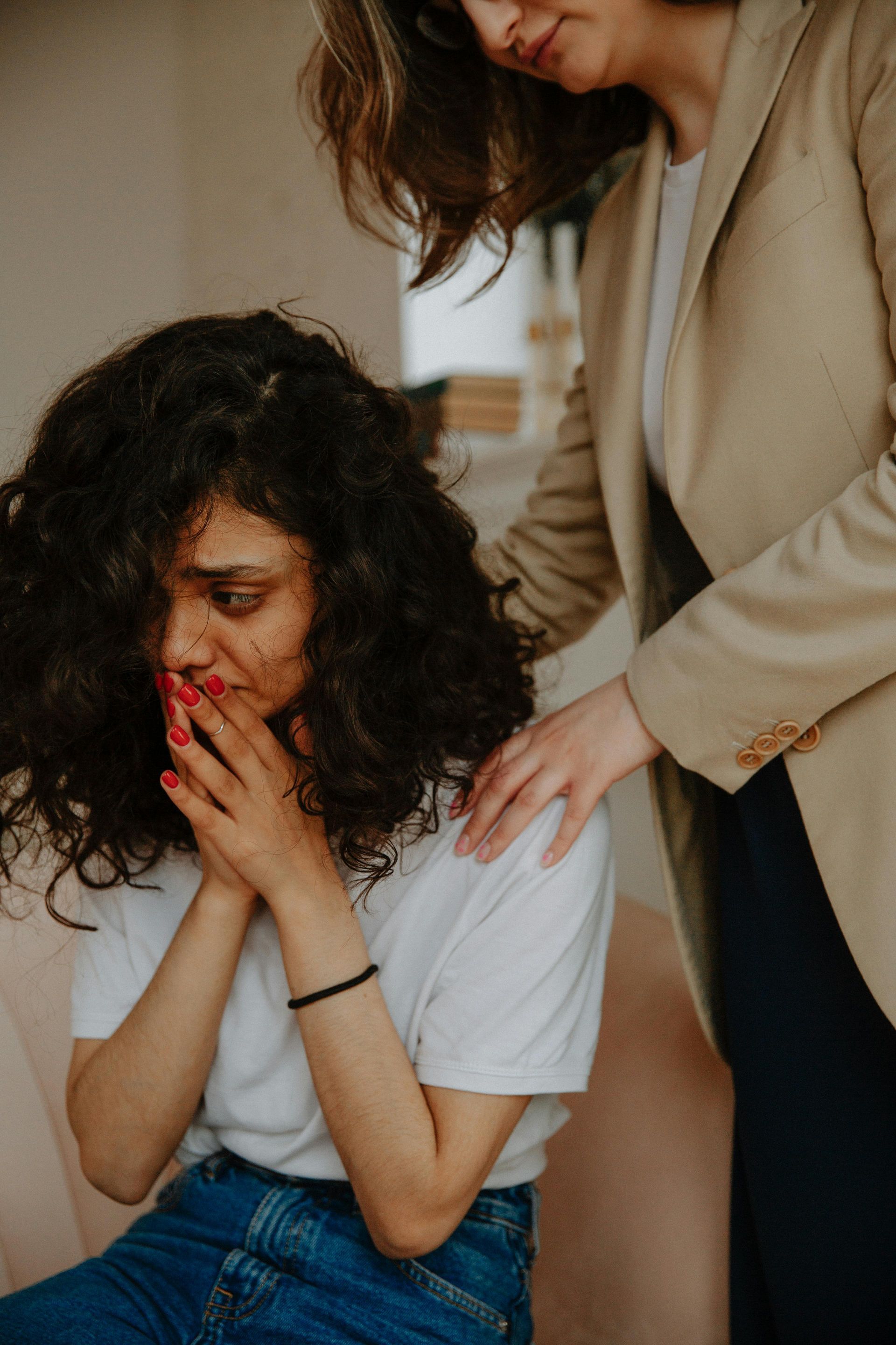 Woman comforts a crying person seated indoors, with a hand on their shoulder.