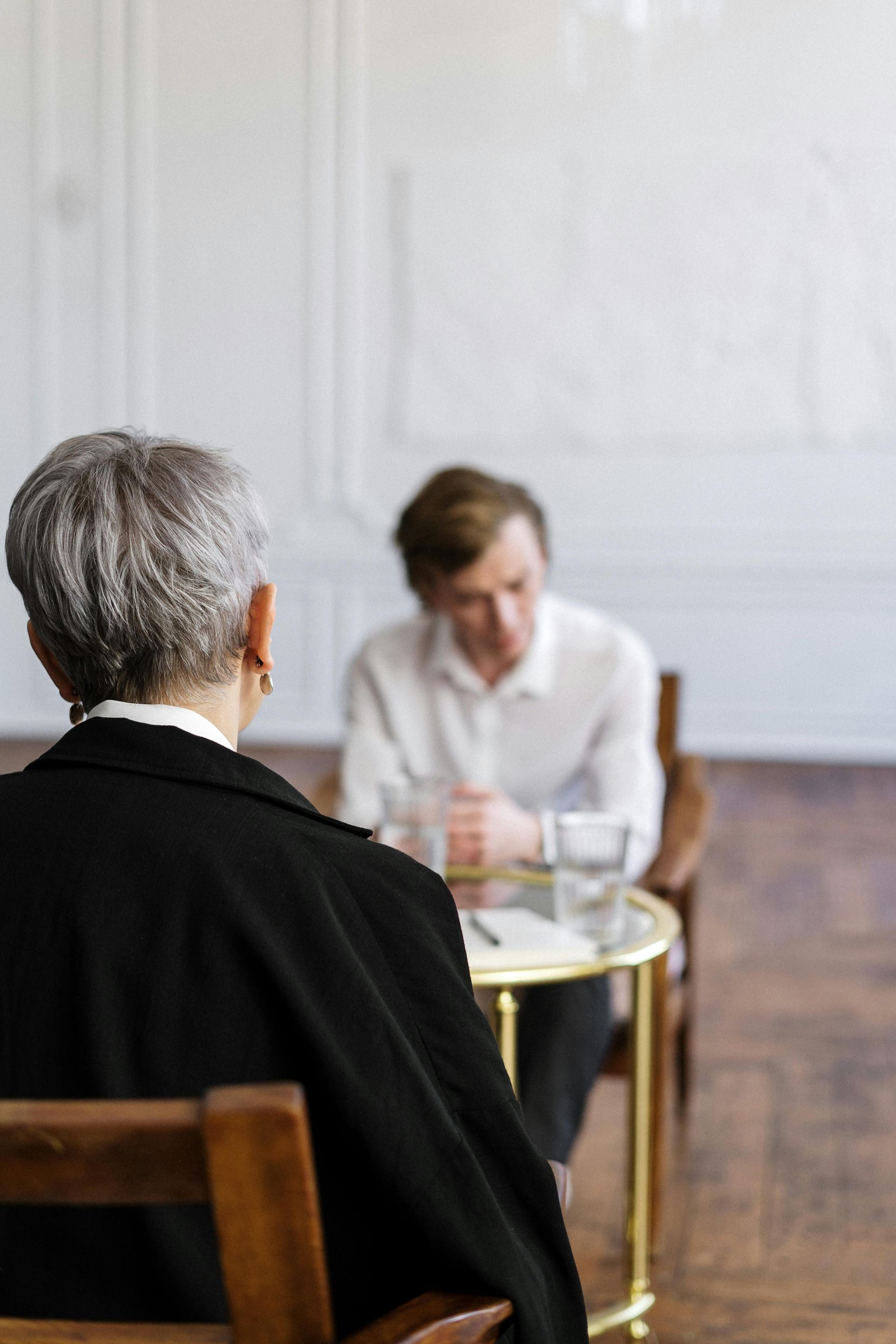 Two people seated across a small table in a bright room, one seen from behind and one leaning forward.