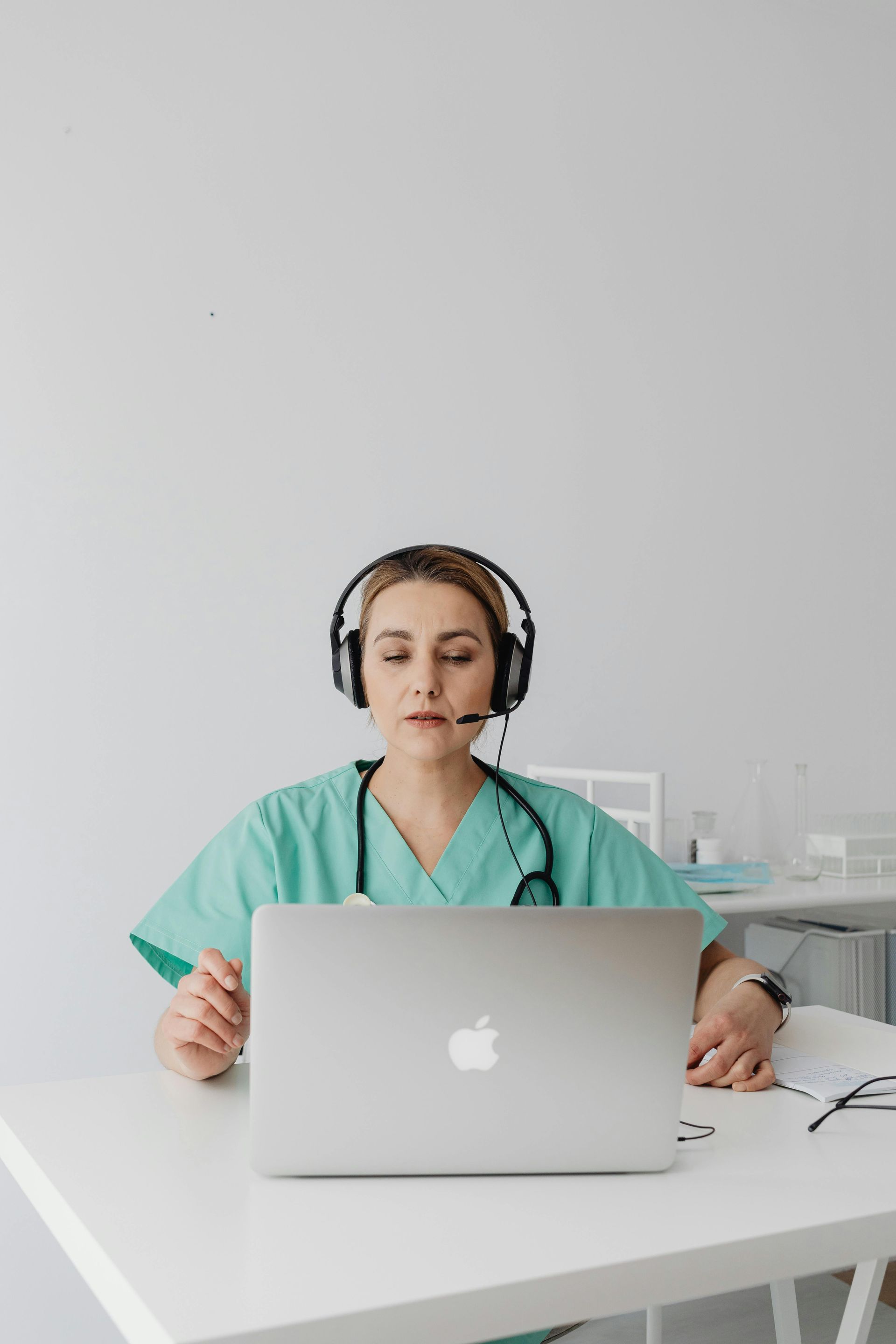 Healthcare worker in teal scrubs wearing headphones, working on a laptop at a white desk