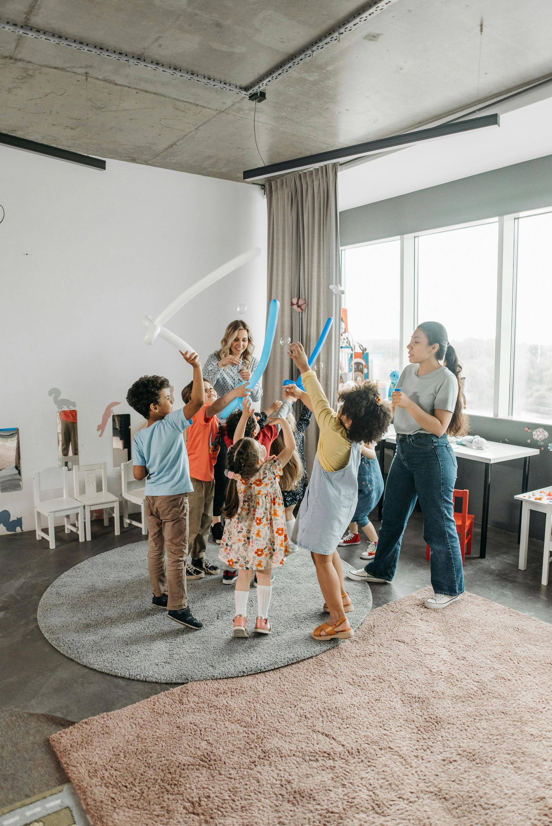Children playing with colorful ribbons in a bright room, with an adult nearby and large windows.