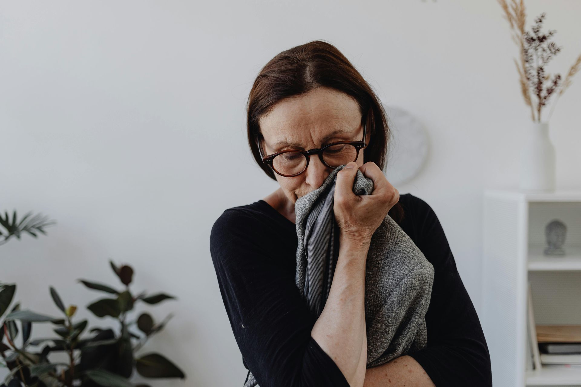 Person wiping tears with a gray cloth, standing indoors beside a plant and shelves