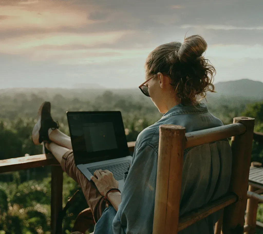 Person relaxing in a wooden chair outdoors, using a laptop with scenic mountain views in the background.