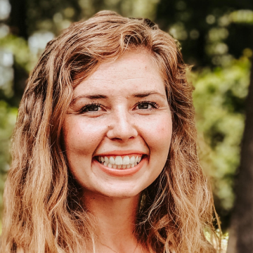 Smiling woman with wavy blonde hair outdoors, with green foliage in the background