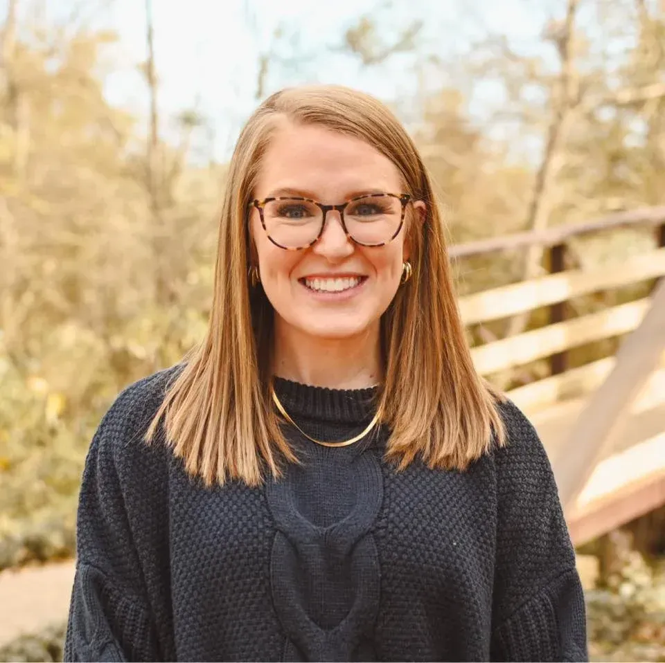 Smiling person with glasses in a black sweater standing outdoors near a wooden railing