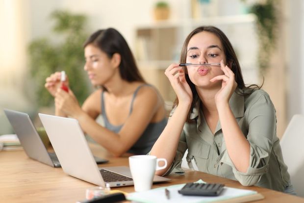 Two women at a table with laptops, one applying lipstick and the other holding a makeup brush