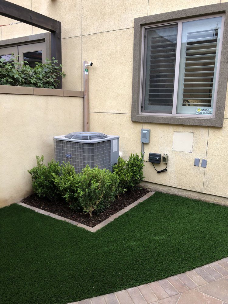 An air conditioner is sitting in a corner of a yard next to a window.