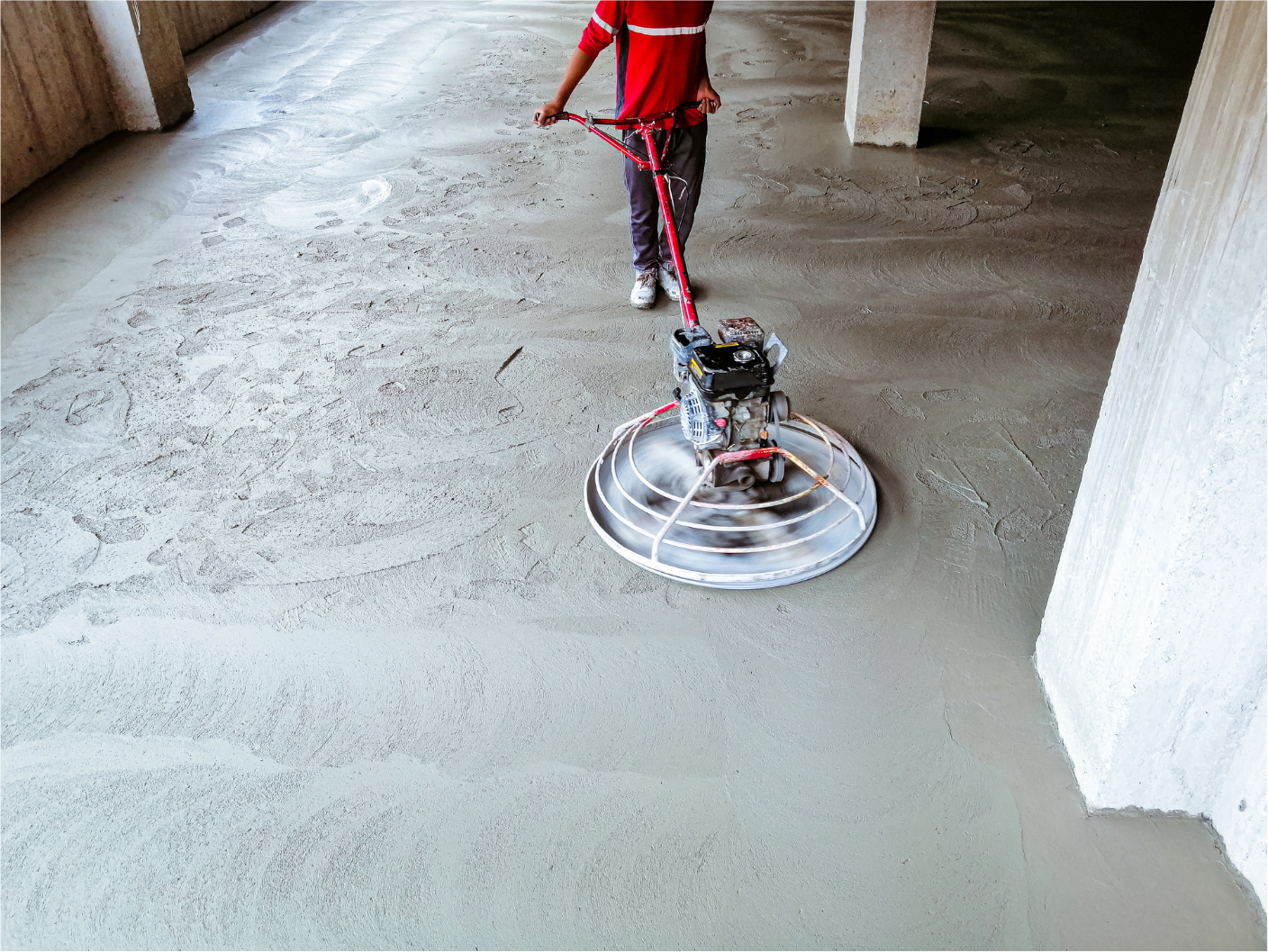 Person using a power trowel on a concrete floor in a building. The man is wearing a red shirt.
