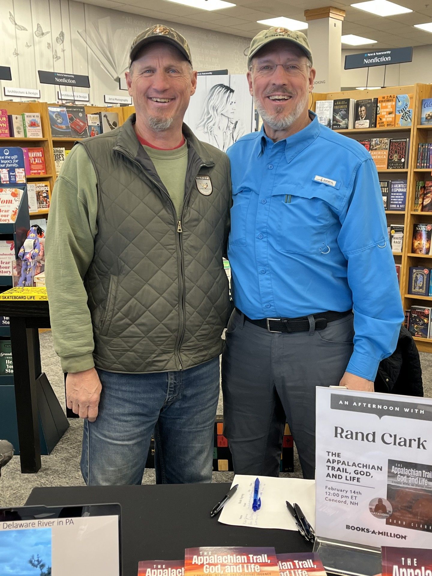 Two men at a book signing table, smiling. One wears a green vest, the other a blue shirt. Bookshelves in the background.