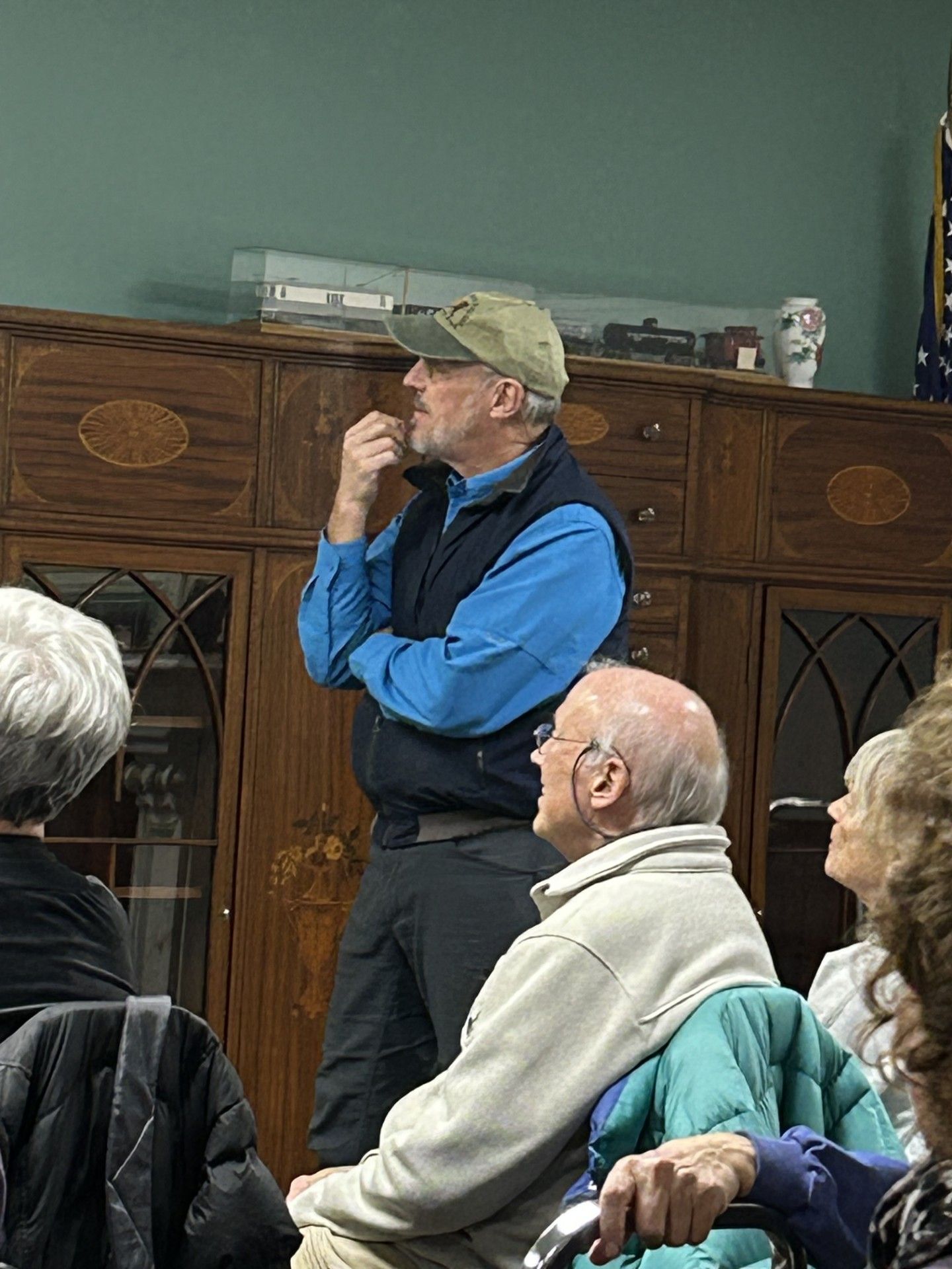 Man speaking at an event, wearing a baseball cap and blue shirt, in front of wooden cabinetry. People seated.