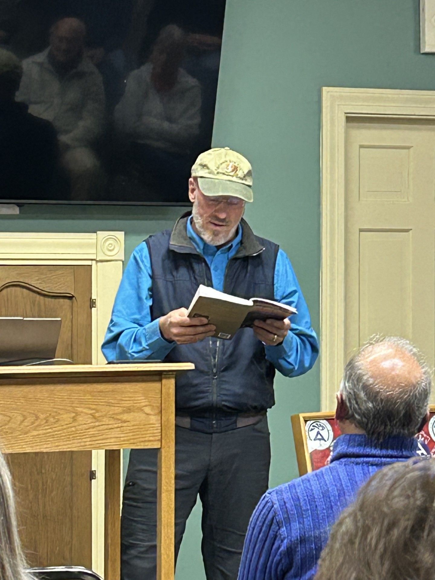 Man reading from a book at a lectern, blue shirt, navy vest, tan cap. People watch.