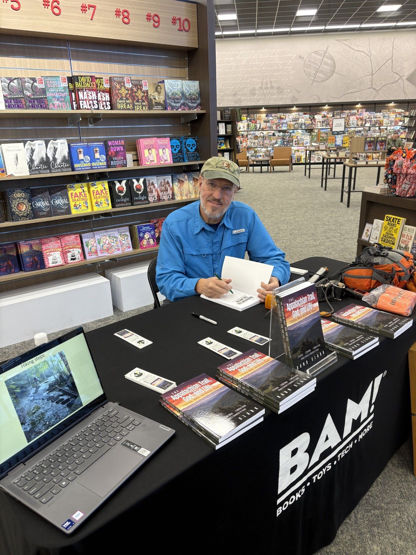 Man at a table signing books in a bookstore. Books on the table, laptop, and shelves of books in the background.