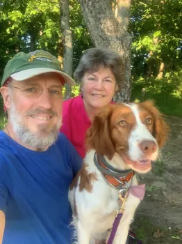 Man and woman smile with a Brittany Spaniel. They're outside by a tree; the dog wears a collar and leash.