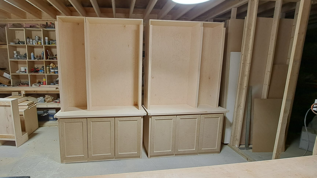 Stacked unfinished wooden cabinets in a workshop, with shelves and lumber in the background
