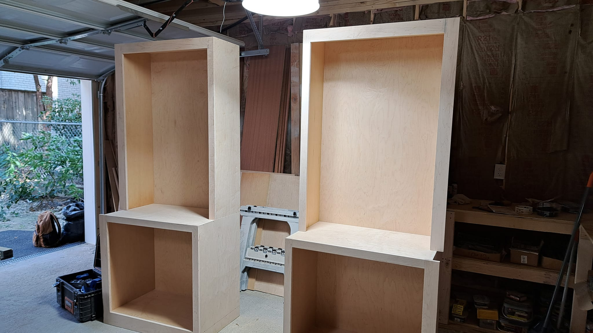 Two unfinished wooden cabinets with open shelves in a workshop, one white and one natural wood.