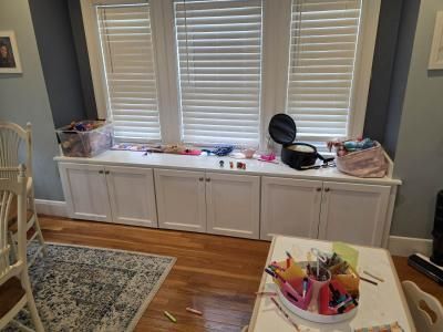 Living room window seat with toys and craft supplies, white cabinets, and hardwood floor
