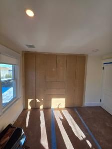Empty sunlit room with built-in wooden cabinets, brown carpet, and a window on the left.