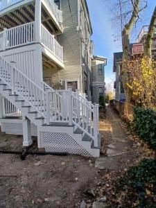 Side view of a white house with stairs and balcony beside a narrow dirt path between buildings