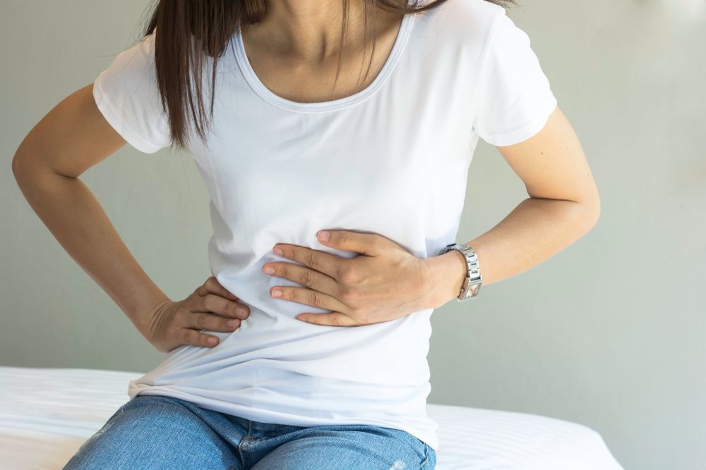Person Sitting on a Bed, Holding Their Stomach With Both Hands — HealthHub Taree in Taree, NSW