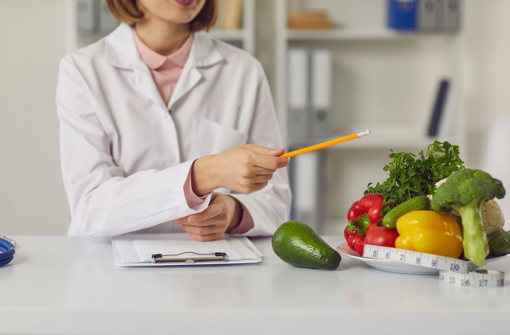 Nutritionist in White Coat Pointing at Fresh Vegetables and Fruit on a Table — HealthHub Taree in Taree, NSW