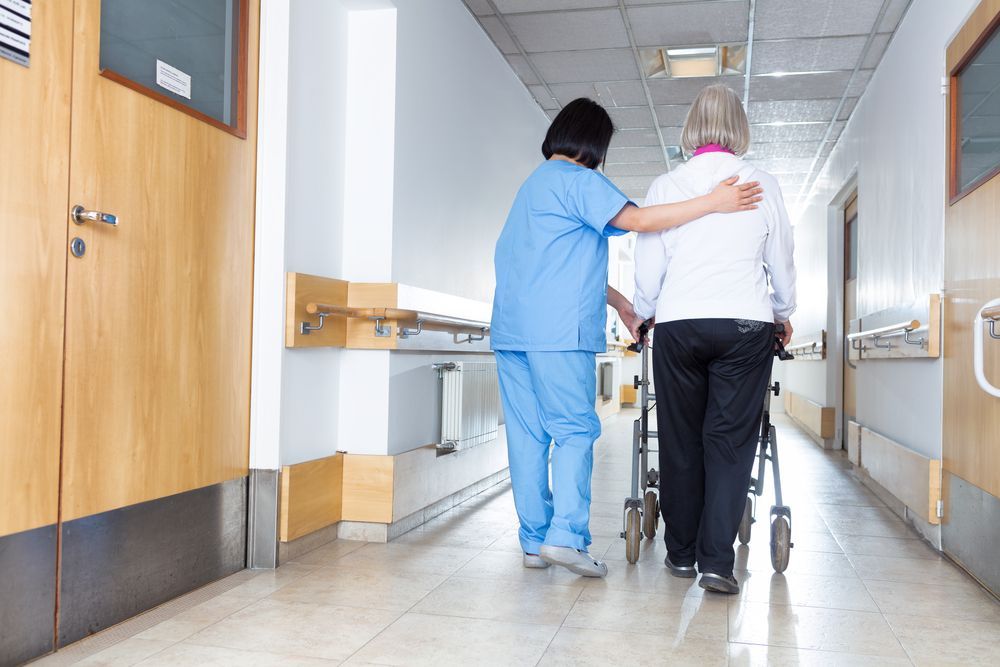 Nurse in Blue Uniform Assisting an Elderly Patient With a Walker Down a Hospital Hallway — HealthHub Taree in Taree, NSW
