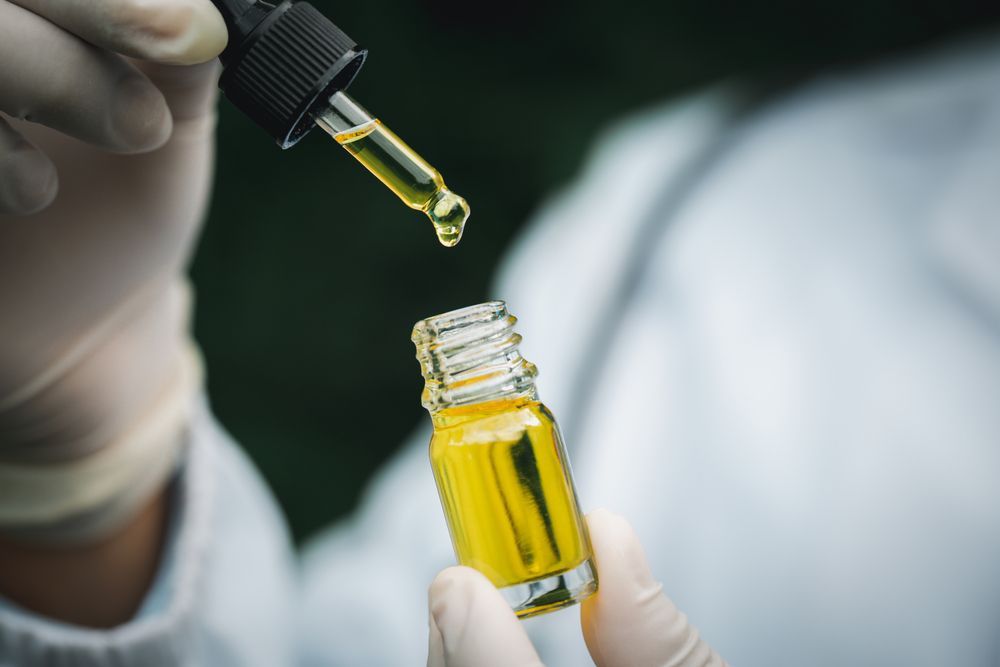 Gloved Hands Holding a Dropper Above a Small Bottle of Yellow Liquid — HealthHub Taree in Taree, NSW