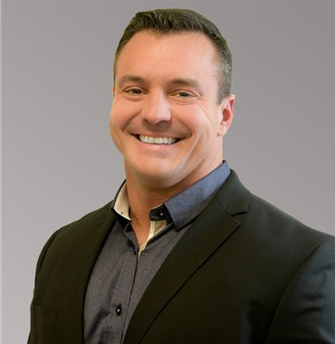 Man smiling, wearing a dark blazer over a blue button-up shirt; neutral background.