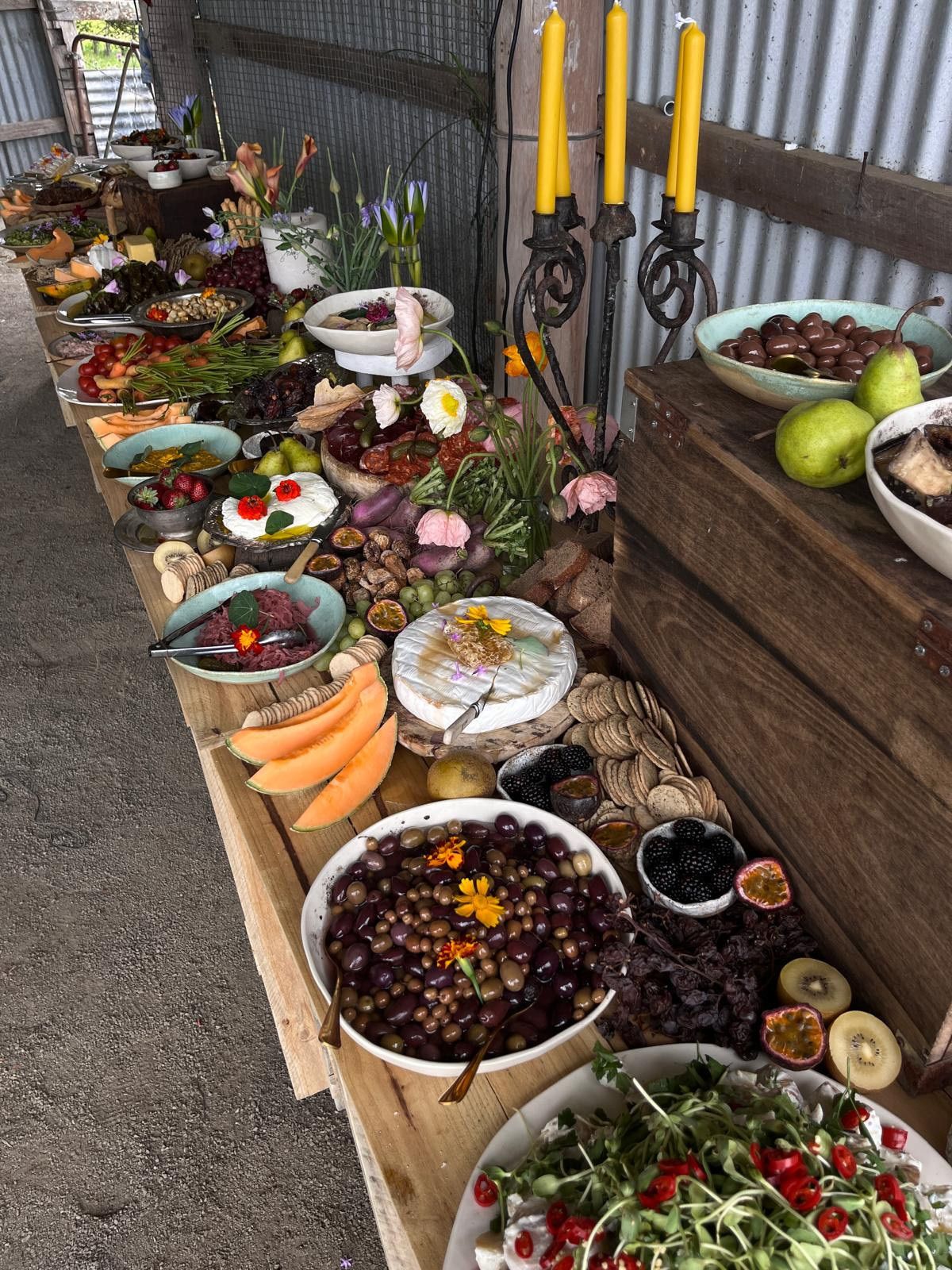 A long wooden table displays an elaborate spread of food: cheeses, fruits, olives, and greens, with candles. — The Habibiz Catering In Wingham, NSW