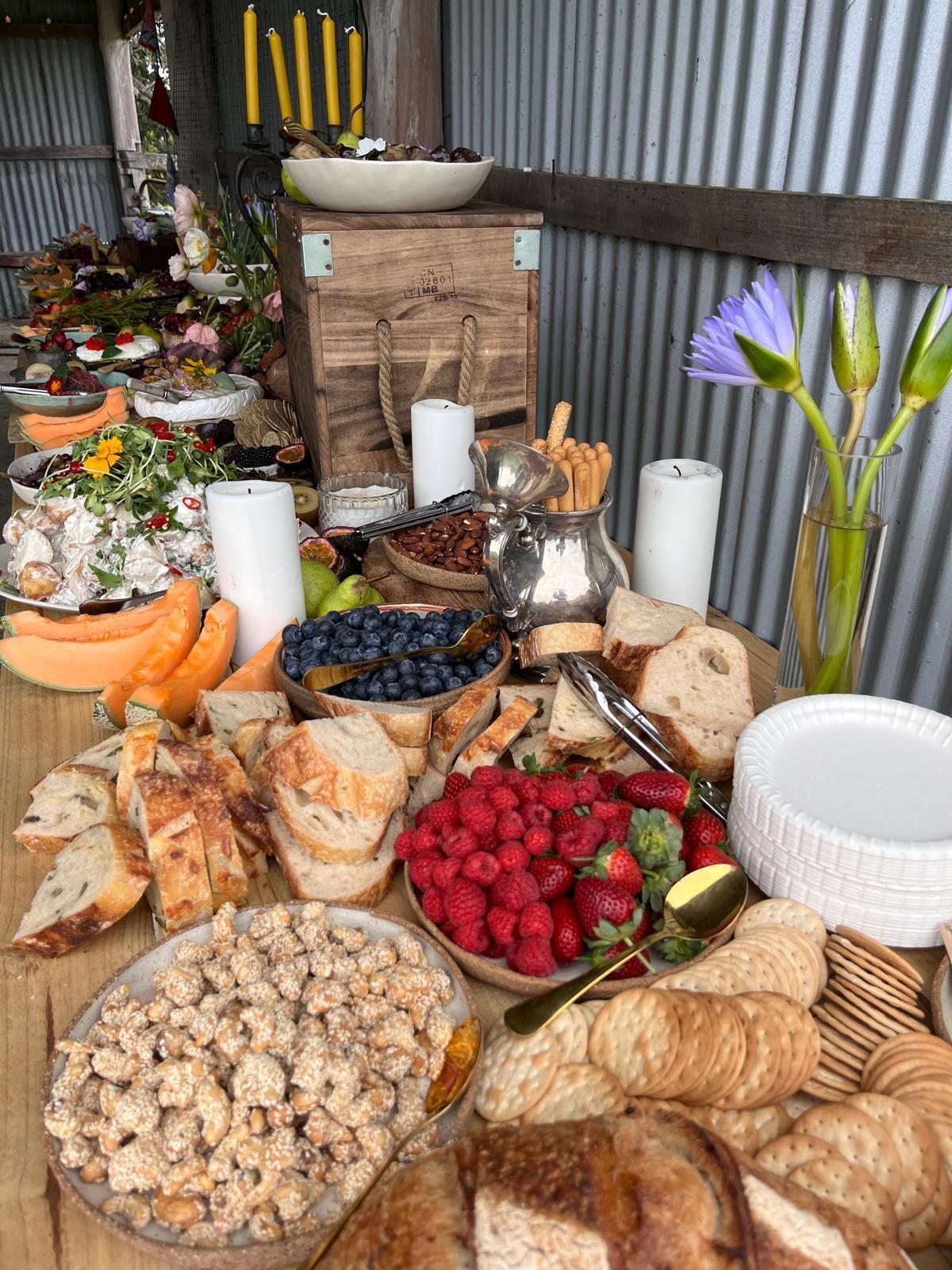 Wooden table laden with food: fruit, bread, pastries, and snacks. A rustic, outdoor setting with a flower vase. — The Habibiz Catering In Wingham, NSW