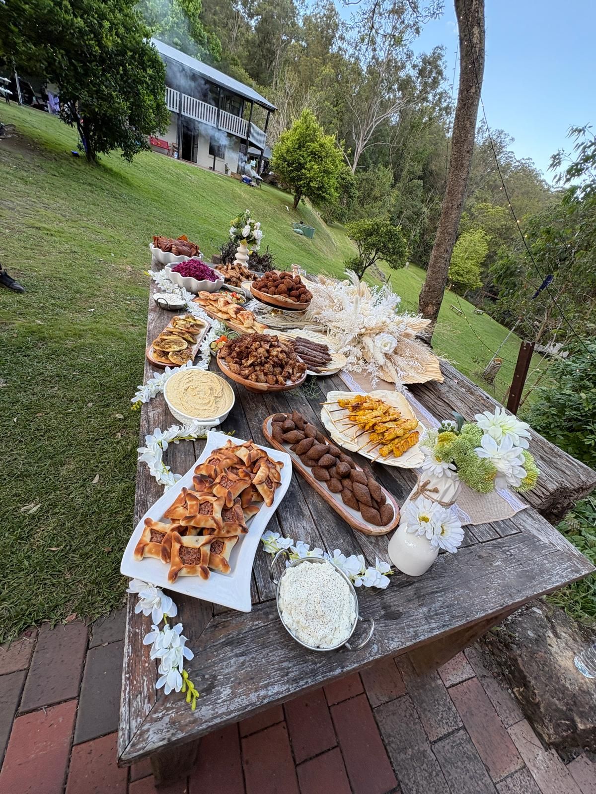 A long wooden table laden with various dishes of food, outdoors on a grassy hillside. — The Habibiz Catering In Wingham, NSW
