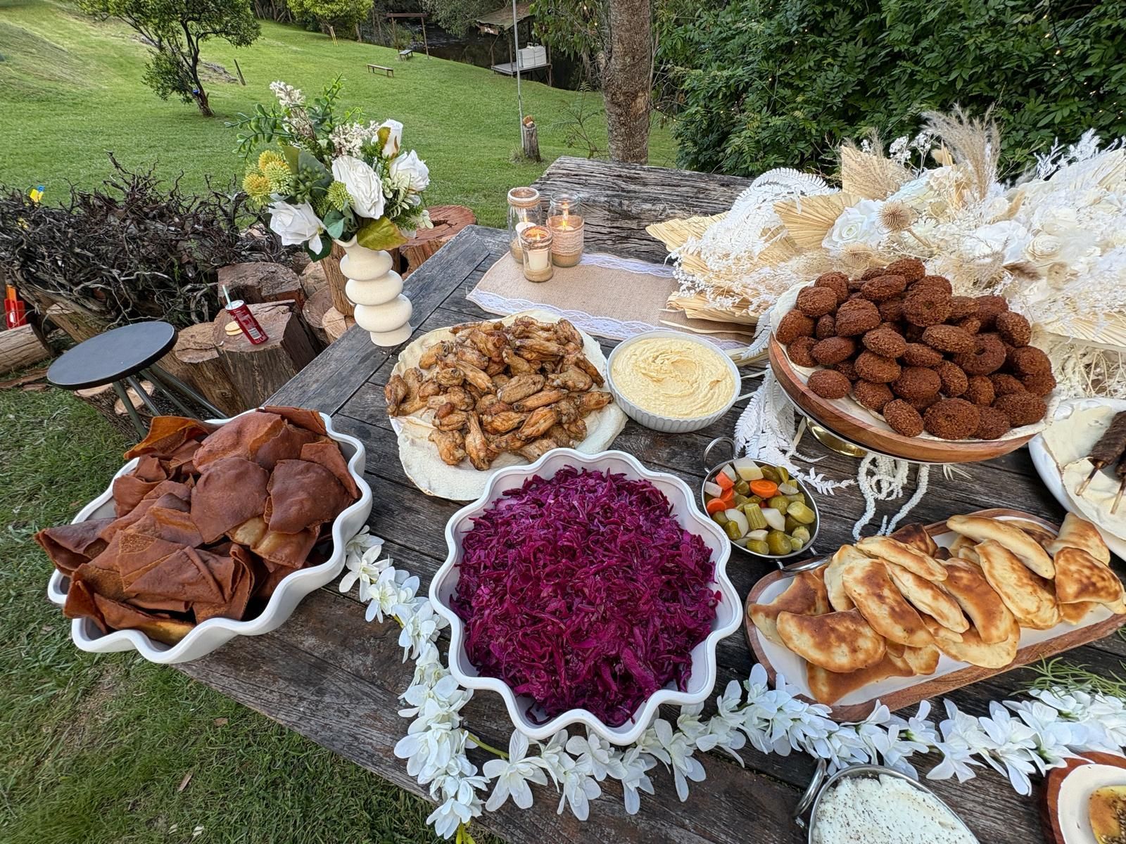 Wooden table laden with various Middle Eastern dishes, including falafel, hummus, and salad. Outdoors with floral decor. — The Habibiz Catering In Wingham, NSW
