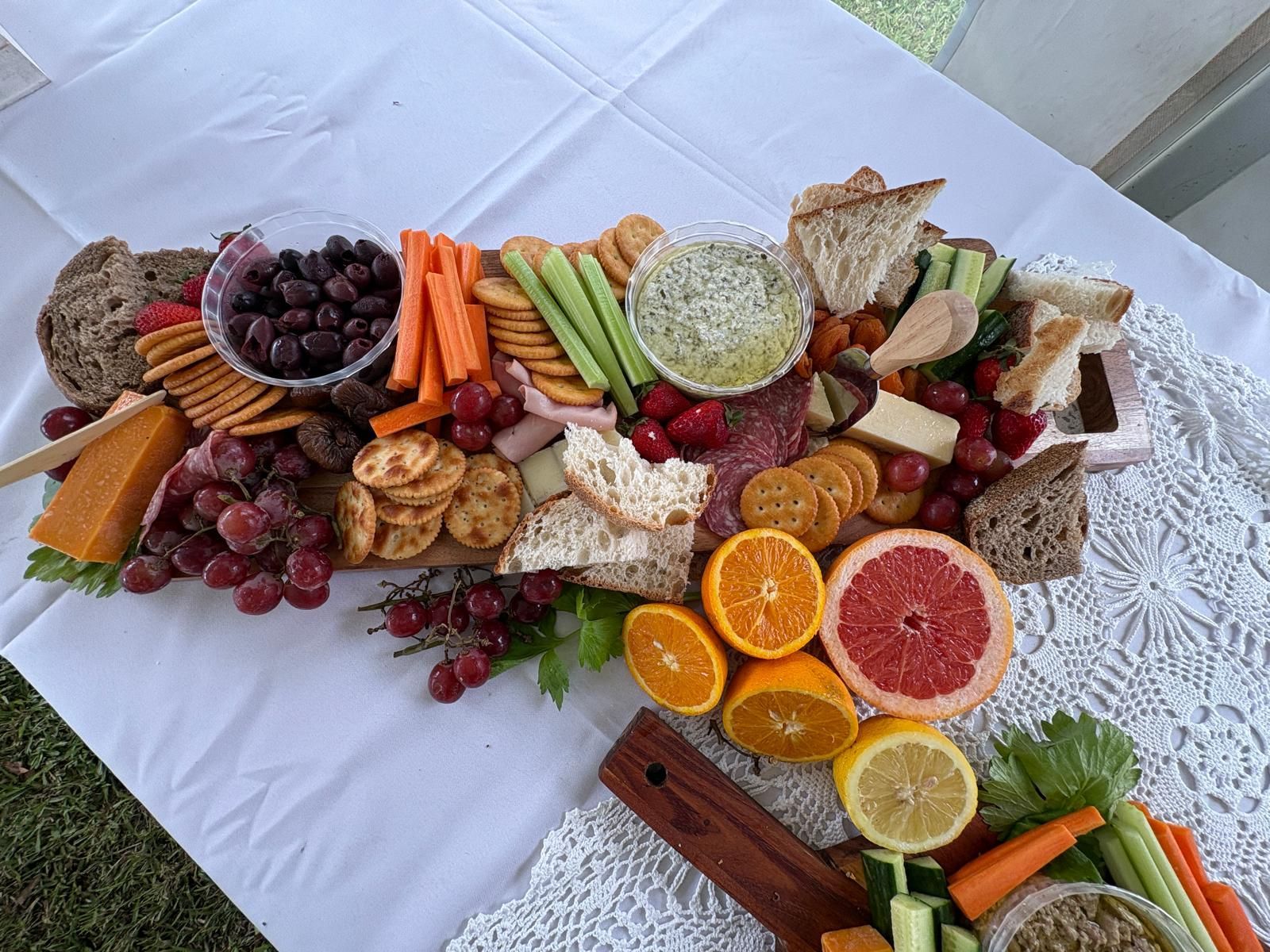Charcuterie board: various foods like fruits, crackers, cheese, and dips arranged on a wooden board on a white tablecloth. — The Habibiz Catering In Wingham, NSW