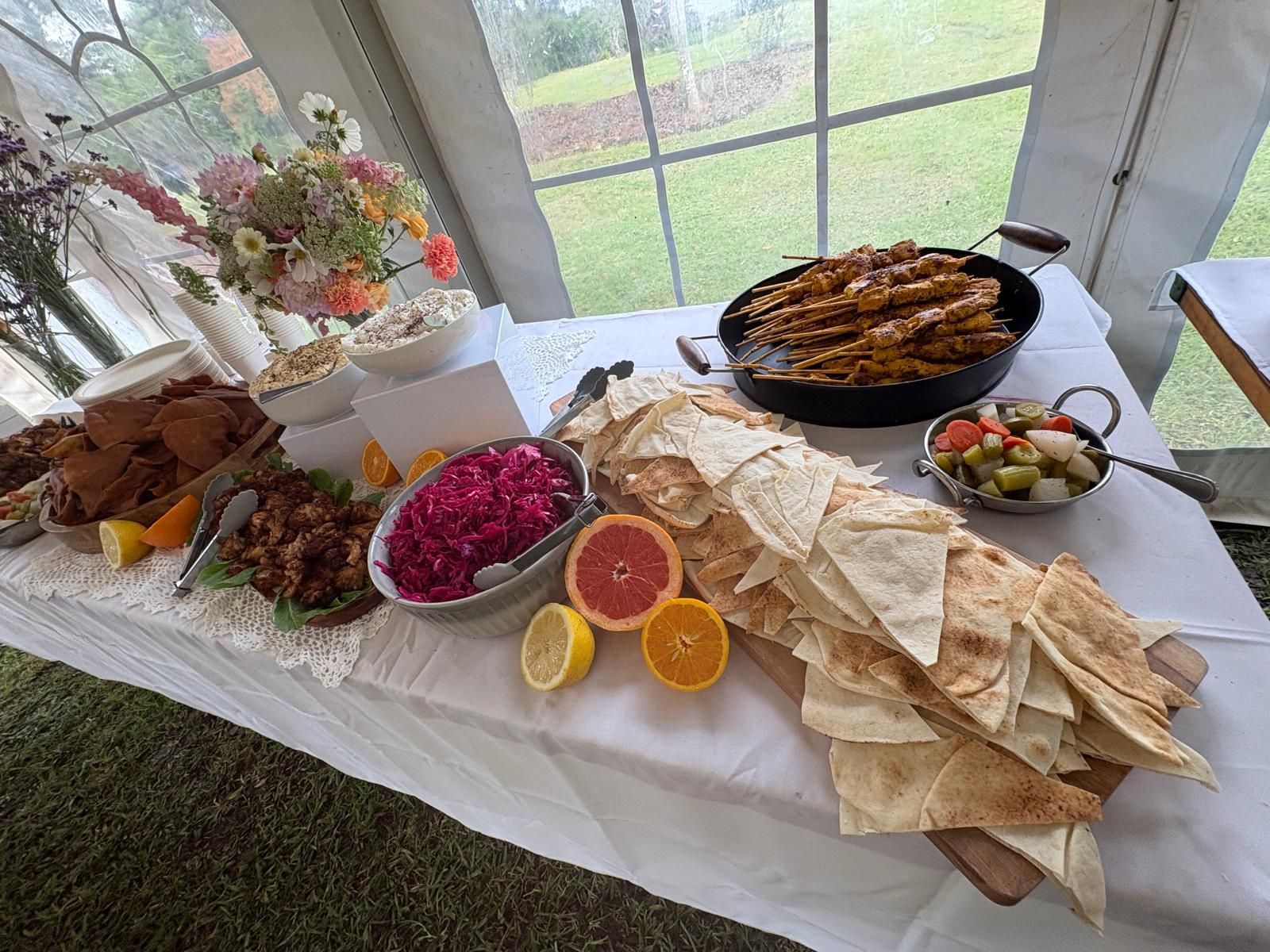 Food buffet on a table with a white cloth, inside a tent. Includes bread, meat skewers, dips, fruit and flowers. — The Habibiz Catering In Wingham, NSW