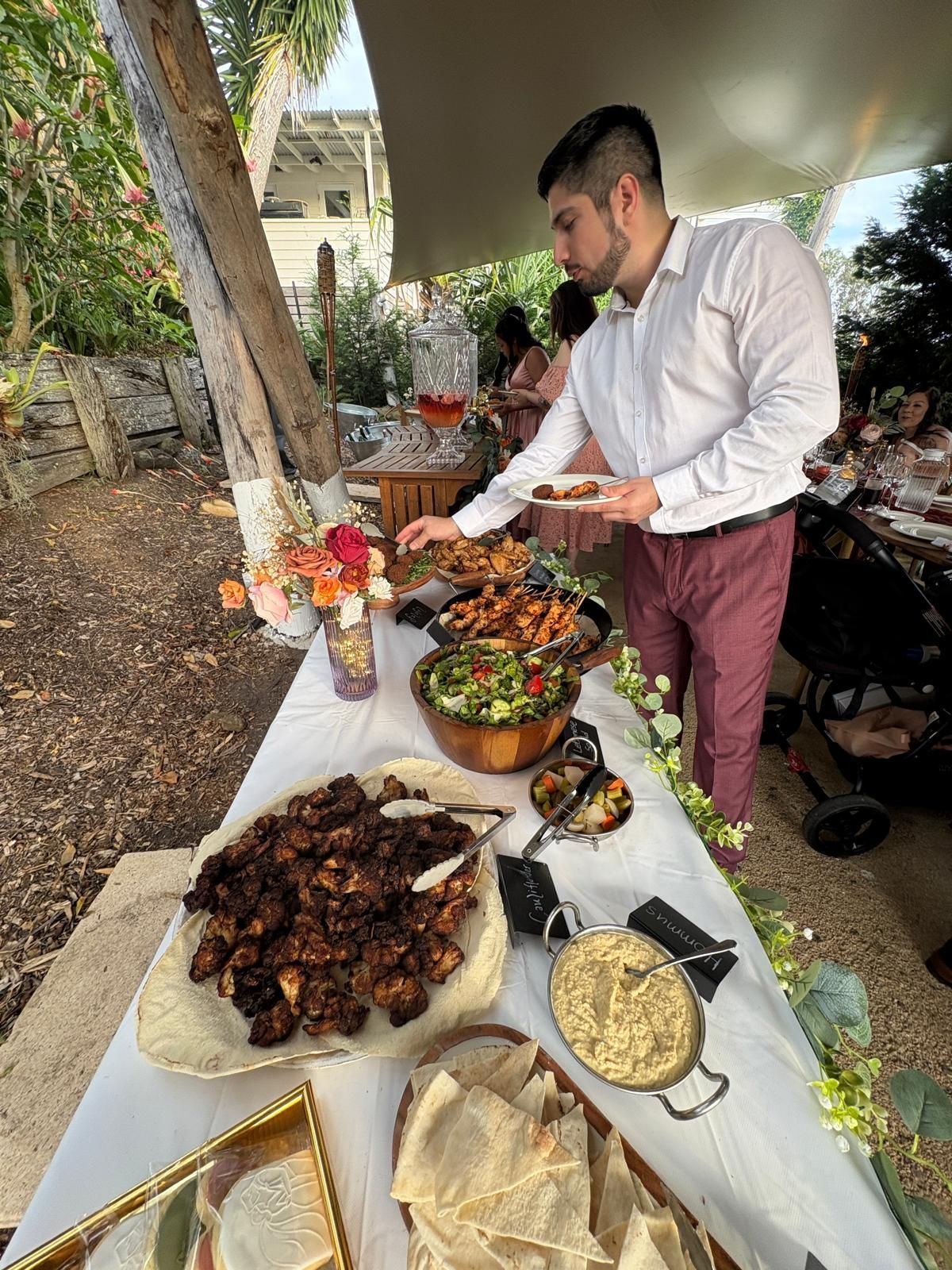 Man serving food from a buffet table at an outdoor event, featuring various dishes and a drink dispenser. — The Habibiz Catering In Wingham, NSW